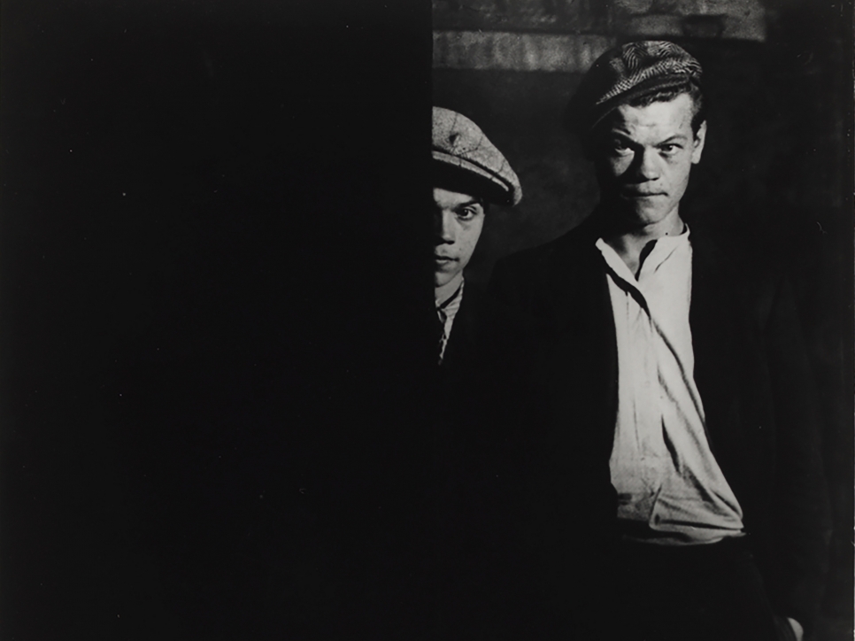 Black and white photographic by Brassaï featuring two men wearing hats. Half of one man's face is showing behind a barrier, and the other's full face is showing 